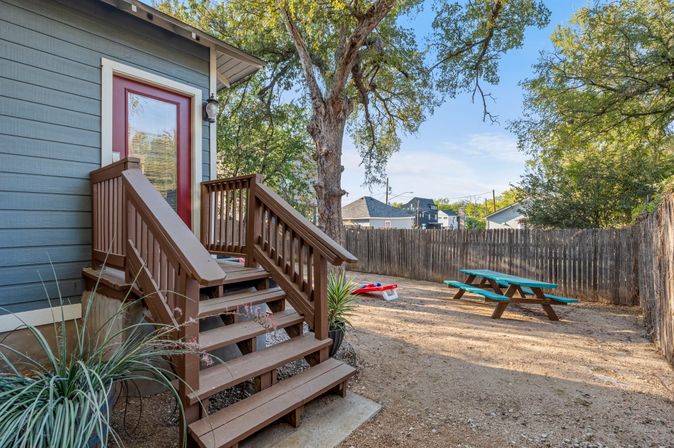 Cozy fenced backyard with brown wooden steps leading to a gray house with a red door, large shade tree, turquoise picnic table and cornhole boards on a gravel yard.