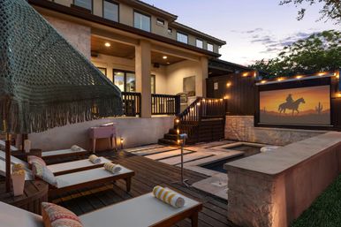 Cozy residential backyard patio at dusk with wooden deck, a narrow plunge pool with stepping stones, multiple lounge chairs with rolled striped towels under a fringed umbrella, warm string lights, and a large outdoor movie screen showing a cowboy silhouette against a desert sunset.