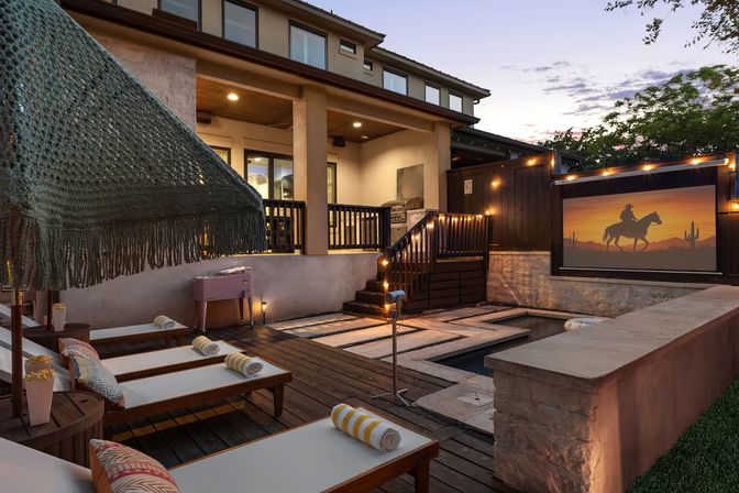 Cozy residential backyard patio at dusk with wooden deck, a narrow plunge pool with stepping stones, multiple lounge chairs with rolled striped towels under a fringed umbrella, warm string lights, and a large outdoor movie screen showing a cowboy silhouette against a desert sunset.