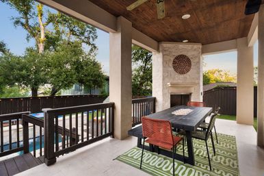 Cozy covered backyard patio with wooden ceiling and stone fireplace, black outdoor dining table with mixed chairs on a green patterned rug, overlooking a pool, deck railing and oak trees.