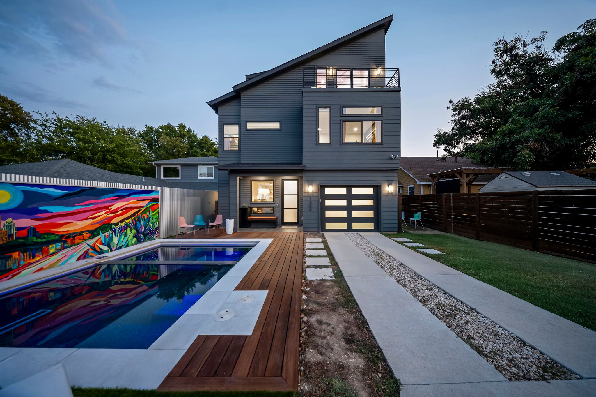 Modern three-story gray home at dusk with illuminated garage, wood-decked backyard pool reflecting a vibrant mural, concrete driveway and patio seating