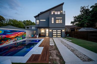 Modern three-story gray home at dusk with illuminated garage, wood-decked backyard pool reflecting a vibrant mural, concrete driveway and patio seating