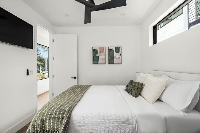 Bright modern minimalist bedroom interior with white walls, wall-mounted TV, black ceiling fan, green knit throw and textured pillows on a white bed, abstract wall art, and a narrow high window with a view of trees.