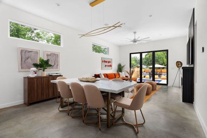 Bright modern open-concept dining and living room with polished concrete floors, marble-top dining table and blush chairs, orange sectional sofa, walnut sideboard, high windows and black-framed sliding doors opening to a sunny backyard patio with pool.