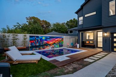 Modern backyard at dusk featuring an illuminated rectangular pool reflecting a vibrant cityscape mural on the fence, wooden deck with white lounge chairs, colorful patio chairs, and a dark-gray two-story house with lit windows.