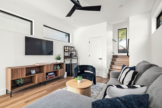 Bright modern living room with wall-mounted TV and walnut media console, gray sectional sofa with black-and-white striped pillows, round wooden coffee table with small potted plant, black accent chair, light hardwood floors, high horizontal windows, black ceiling fan and stairs leading up.