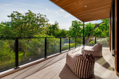 Sunlit covered balcony with glass railing overlooking lush treetops, striped outdoor bean bag chairs on a concrete floor beneath a wood-paneled ceiling — a relaxing patio view.