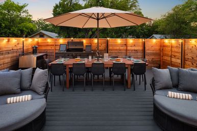 Cozy backyard deck at dusk with a large umbrella over a wooden dining table for eight, gas grill, string lights, and cushioned lounge seating.