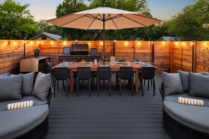 Cozy backyard deck at dusk with a large umbrella over a wooden dining table for eight, gas grill, string lights, and cushioned lounge seating.