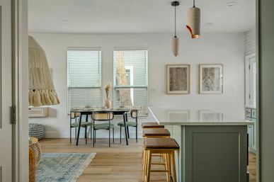 Sunlit coastal-style open kitchen and dining area with white island, three wooden bar stools, pendant lights, rattan-backed dining chairs, large windows and light wood floors.