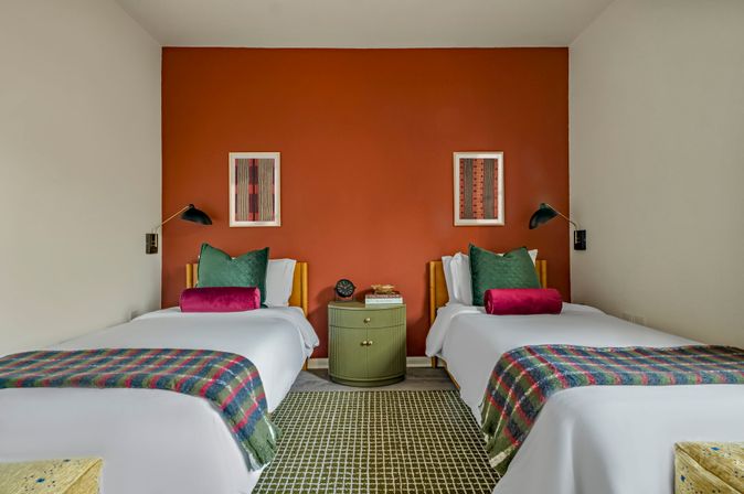Stylish twin-bed hotel room with a rust-orange accent wall, two white beds topped with green pillows and magenta bolster cushions, plaid throws, a round green bedside table with a clock, wall-mounted reading lamps and framed art