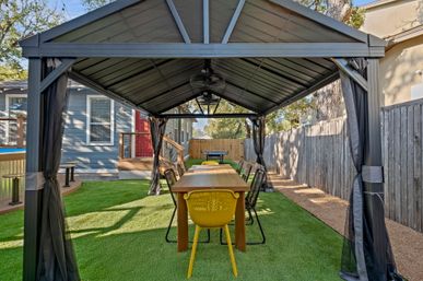 Covered metal gazebo over a long wooden dining table with mixed chairs, a bright yellow chair at the near end, ceiling fans, and artificial turf in a suburban backyard beside a wooden fence and blue house.