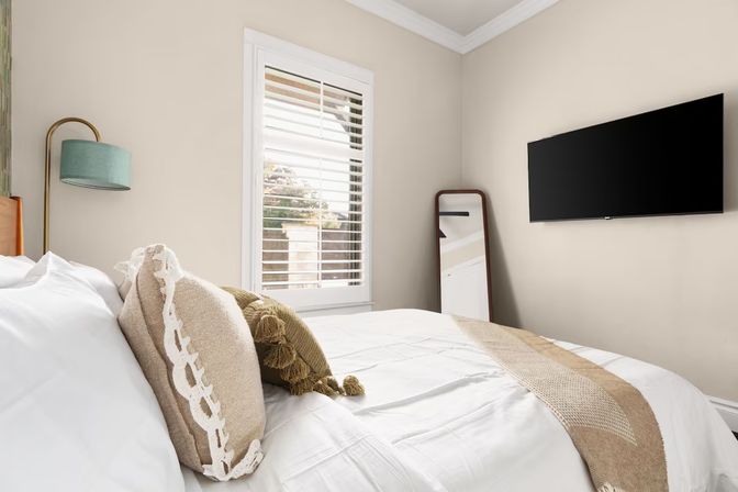 Sunlit modern neutral bedroom with white bedding, textured beige throw pillows and fringe blanket, teal arc floor lamp, window shutters, leaning full-length mirror, and wall-mounted flat-screen TV.