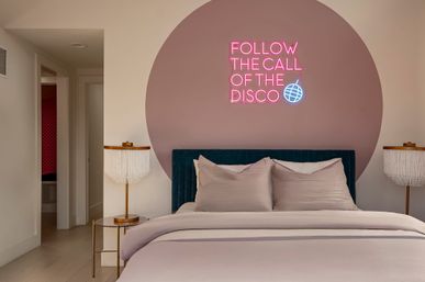 Cozy modern bedroom with a blush circular accent wall and pink neon sign reading FOLLOW THE CALL OF THE DISCO, teal velvet headboard, soft neutral bedding and fringed bedside lamps.