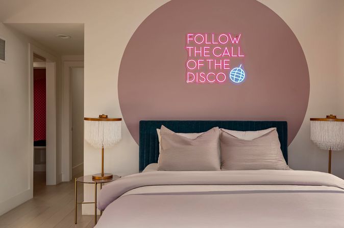 Cozy modern bedroom with a blush circular accent wall and pink neon sign reading FOLLOW THE CALL OF THE DISCO, teal velvet headboard, soft neutral bedding and fringed bedside lamps.