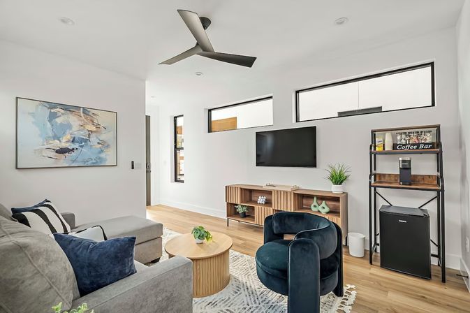 Bright modern living room with light wood floors, gray sectional and navy pillows, round wood coffee table, deep teal velvet accent chair, wall-mounted TV above a slatted wood media console, small coffee station with mini fridge, abstract art and black ceiling fan