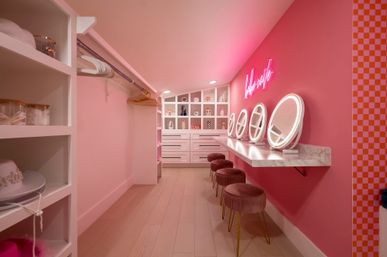 Playful pink walk-in dressing room with marble countertop vanity, four round lighted mirrors, velvet stools, open shelving and drawers, neon wall sign and wood plank floor — boutique-style makeup station.
