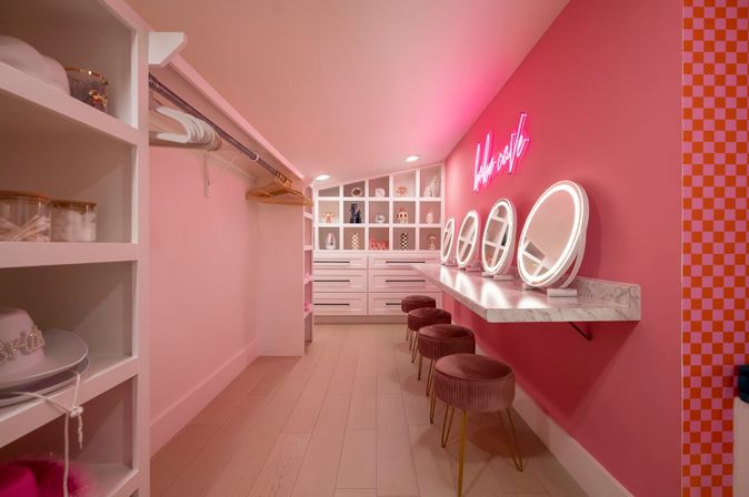 Playful pink walk-in dressing room with marble countertop vanity, four round lighted mirrors, velvet stools, open shelving and drawers, neon wall sign and wood plank floor — boutique-style makeup station.