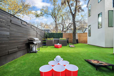 Modern urban backyard with bright artificial turf featuring a red-cup beer-pong setup and white ball in the foreground, cornhole boards, gas grill along a dark wood fence, planter boxes and patio seating beside a white multi-story house under trees.