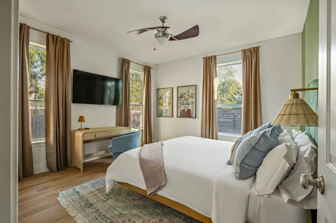 Sunlit modern bedroom with a queen bed dressed in layered pillows, wall-mounted TV, wooden desk and blue accent chair, ceiling fan, hardwood floors and neutral curtains