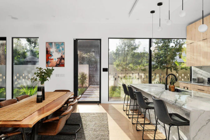 Bright modern open-plan kitchen and dining room with floor-to-ceiling windows facing a leafy backyard, marble island with black bar stools, live-edge wood dining table with leather chairs, pendant lights and natural light-filled contemporary interior.