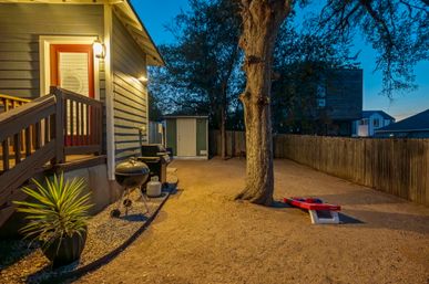 Twilight backyard with gravel ground, large oak tree, wooden privacy fence, lit deck entrance, charcoal and gas grills beside a potted plant, storage shed, and a red cornhole board for outdoor games.