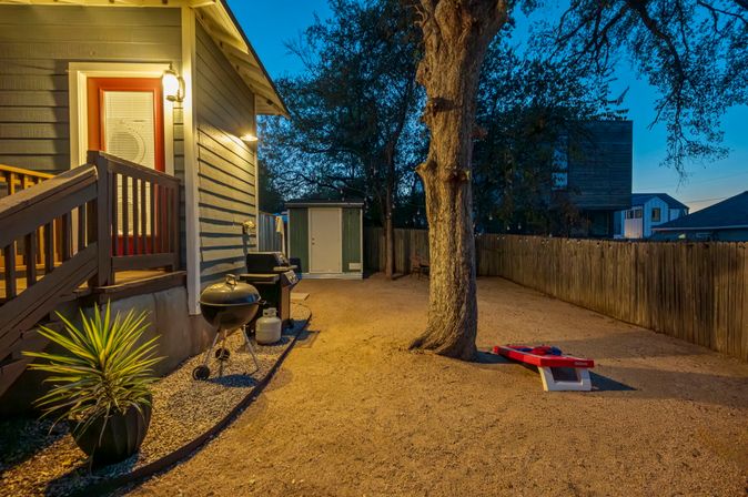 Twilight backyard with gravel ground, large oak tree, wooden privacy fence, lit deck entrance, charcoal and gas grills beside a potted plant, storage shed, and a red cornhole board for outdoor games.