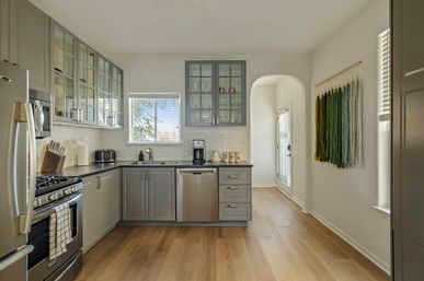 Sunlit contemporary gray kitchen with stainless-steel refrigerator, gas range and dishwasher, glass-front upper cabinets, white subway tile backsplash, light hardwood floors, arched doorway and green ombré yarn wall hanging