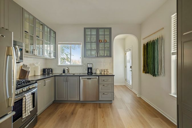 Sunlit contemporary gray kitchen with stainless-steel refrigerator, gas range and dishwasher, glass-front upper cabinets, white subway tile backsplash, light hardwood floors, arched doorway and green ombré yarn wall hanging