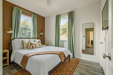 Cozy bedroom with queen bed, brown accent wall, two tall windows with green curtains, brass wall sconces, rattan nightstands, patterned throw pillows, full‑length mirror and wood‑look flooring.