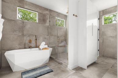 Contemporary bathroom with geometric freestanding bathtub, glass walk-in shower, large gray stone tiles, brass fixtures, rolled towels and clerestory windows showing green trees.