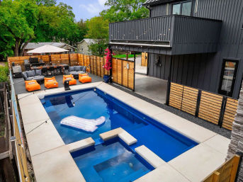 Modern backyard with a rectangular blue swimming pool and attached spa, white inflatable float on the water, concrete deck, adjacent patio with gray sofas and bright orange beanbag loungers, dining table under an umbrella, wood privacy fence, string lights and a black contemporary house exterior.