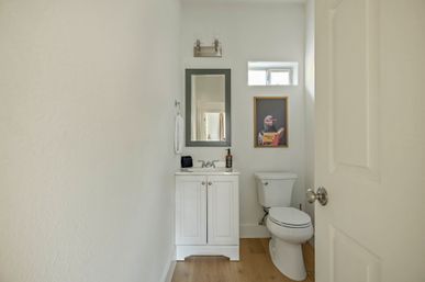Small modern half-bath with white vanity and mirror, toilet on light wood floor, small high window and framed playful monkey art wearing glasses
