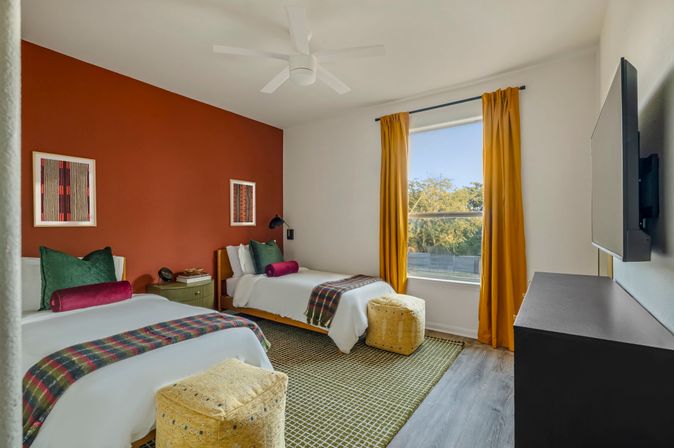 Sunlit modern twin bedroom with rust accent wall, mustard curtains, plaid throws, green pillows and textured yellow poufs