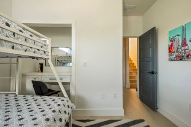 Bright modern residential bedroom with white bunk bed and sunburst-patterned bedding, built-in white desk and black chair, gray interior door open to staircase, striped rug and colorful cityscape wall art.