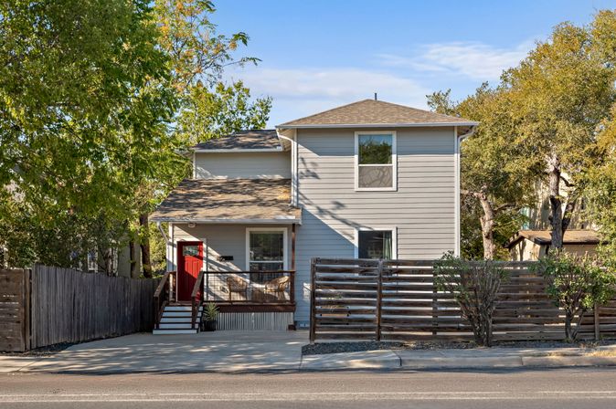 Two-story gray house with an inviting red front door, small covered porch, wood-slat privacy fence and driveway on a tree-lined suburban street