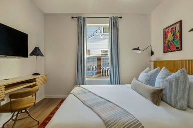 Cozy modern guest bedroom with white linens and striped throw, wooden headboard, three blue-striped pillows and lumbar cushion, wall-mounted reading lamp, wall TV and slim wooden desk with vintage chair, window framed by blue curtains with outdoor string lights visible