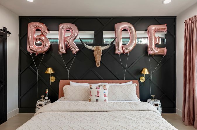 Modern bedroom with black geometric accent wall and rose-gold foil balloons spelling "BRIDE" over a blush upholstered bed, decorative longhorn skull, gold wall sconces, patterned white bedding and a western boot-print pillow