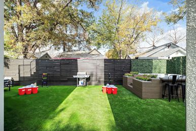 Sunny suburban backyard with bright green artificial turf, dark horizontal privacy fence, gas grill, raised wooden planter boxes with bar seating, and red-cup yard game setup under leafy trees.