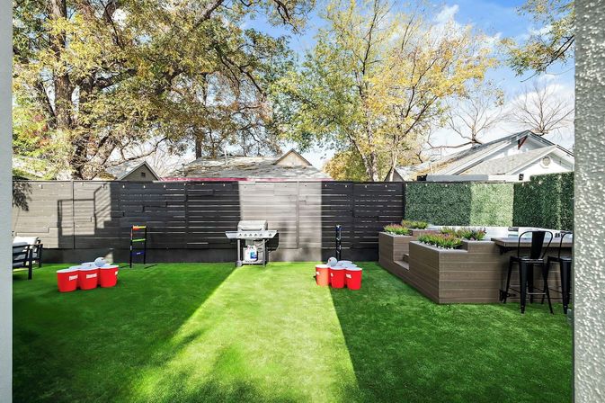 Sunny suburban backyard with bright green artificial turf, dark horizontal privacy fence, gas grill, raised wooden planter boxes with bar seating, and red-cup yard game setup under leafy trees.