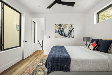 Bright modern bedroom with white walls and hardwood floors, large windows and black ceiling fan, queen bed with white linens and navy throw, patterned rug, abstract wall art, bedside lamp and open door to stairwell.