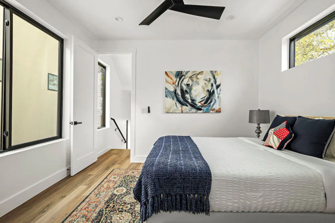 Bright modern bedroom with white walls and hardwood floors, large windows and black ceiling fan, queen bed with white linens and navy throw, patterned rug, abstract wall art, bedside lamp and open door to stairwell.