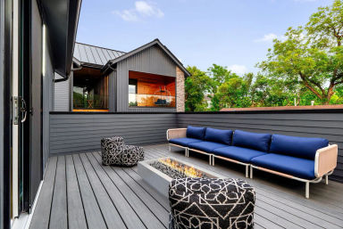Contemporary rooftop deck with gray composite decking and siding, long navy-cushioned outdoor sofa, black-and-white patterned poufs, low linear fire pit, and leafy suburban trees beyond.