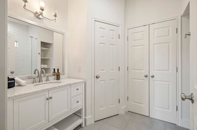 Bright modern white bathroom with marble-topped vanity, chrome faucet, large mirror, tiled floor and double closet doors.
