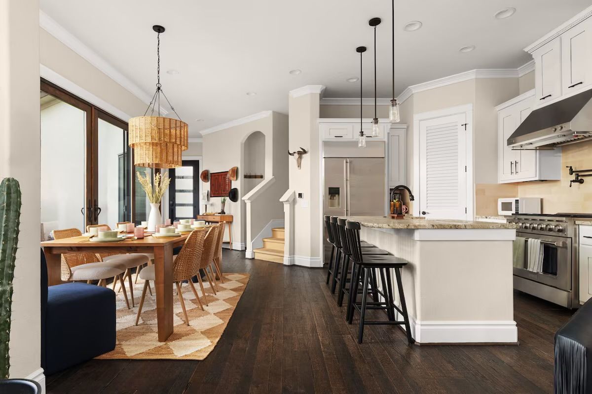 Sunlit open-plan kitchen and dining area in a modern family home — dark hardwood floors, wood dining table with wicker chairs under a woven rattan pendant, sliding glass doors, kitchen island with granite countertop and black bar stools, stainless steel appliances.