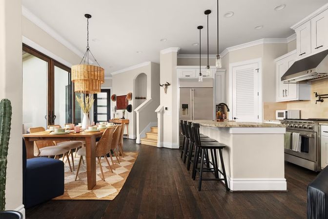 Sunlit open-plan kitchen and dining area in a modern family home — dark hardwood floors, wood dining table with wicker chairs under a woven rattan pendant, sliding glass doors, kitchen island with granite countertop and black bar stools, stainless steel appliances.