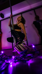 Pole dancer in black outfit and chrome platform heels posing on a vertical pole in a purple-lit studio with scattered purple string lights