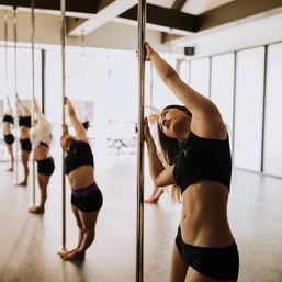 Sunlit pole fitness class in a bright studio, women stretching and practicing moves on vertical poles with a focused participant leaning against a pole in the foreground.