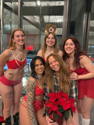 Five smiling women in red holiday outfits posing inside a downtown high-rise by a large window, one wearing reindeer antlers and another holding a poinsettia for a festive group photo.
