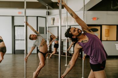 Energetic pole fitness class in a bright dance studio, participants leaning into synchronized side stretches on chrome poles.